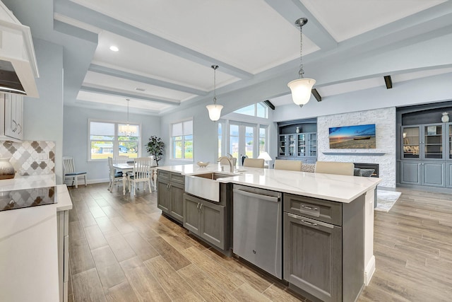 kitchen featuring stainless steel dishwasher, gray cabinetry, decorative light fixtures, a stone fireplace, and an island with sink