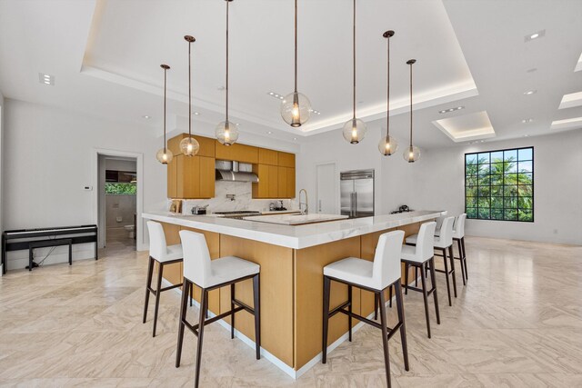 kitchen with wall chimney exhaust hood, decorative light fixtures, a raised ceiling, a kitchen bar, and built in fridge