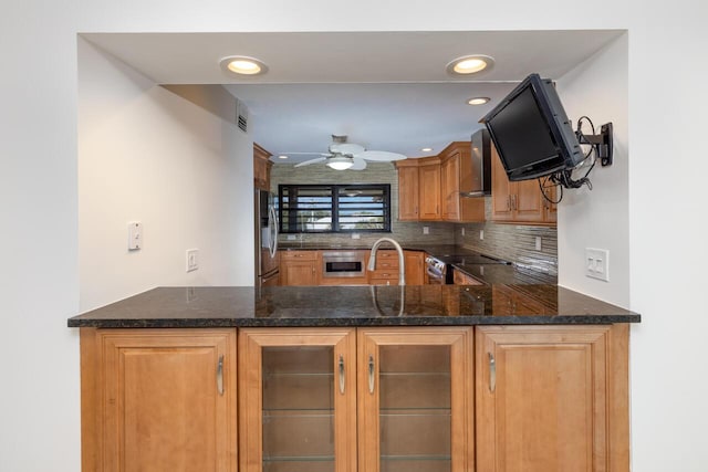 kitchen with ceiling fan, wall chimney exhaust hood, tasteful backsplash, dark stone counters, and appliances with stainless steel finishes