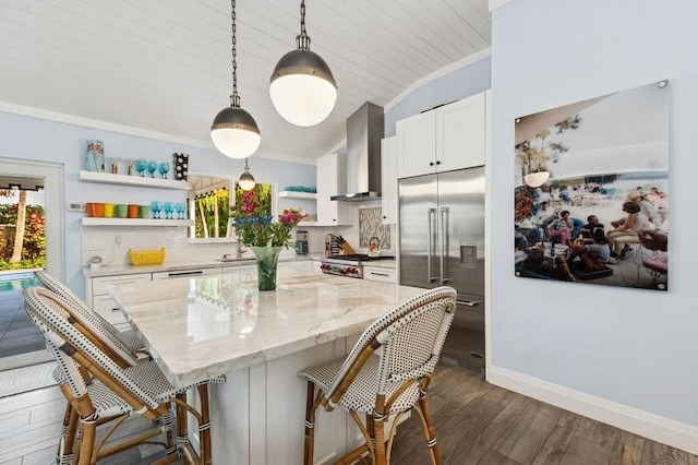 kitchen featuring pendant lighting, stainless steel built in refrigerator, wall chimney range hood, light stone counters, and white cabinetry