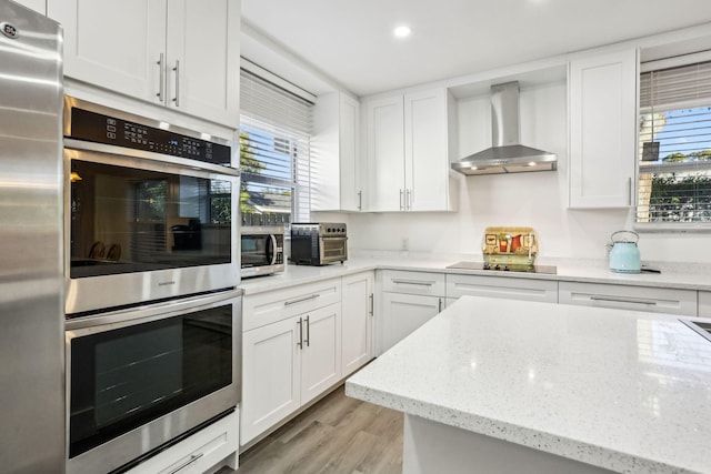 kitchen featuring light stone countertops, wall chimney exhaust hood, light wood-type flooring, white cabinets, and appliances with stainless steel finishes