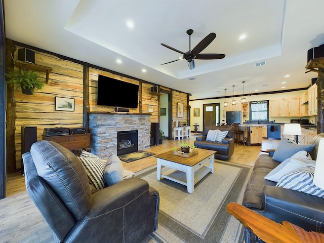 living room featuring a raised ceiling, ceiling fan, light hardwood / wood-style flooring, a fireplace, and wood walls