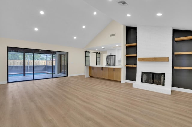 unfurnished living room featuring a barn door, built in features, high vaulted ceiling, a fireplace, and light wood-type flooring