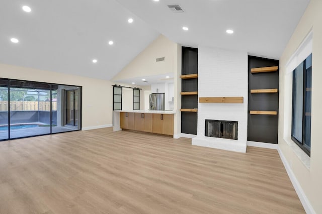 unfurnished living room featuring built in shelves, a barn door, high vaulted ceiling, a fireplace, and light wood-type flooring