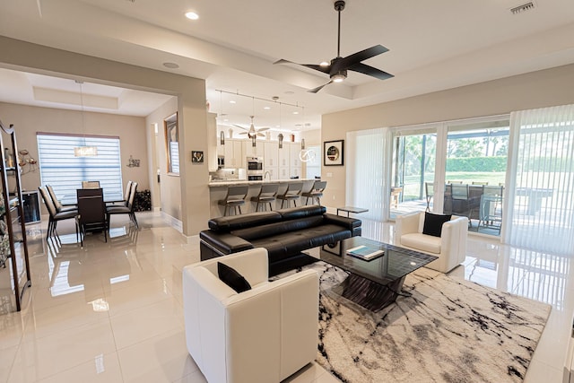 living room featuring light tile patterned floors, ceiling fan with notable chandelier, and a tray ceiling