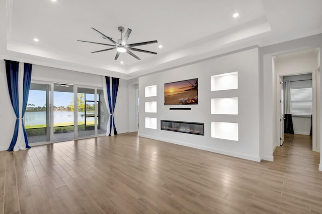 unfurnished living room featuring a tray ceiling, light hardwood / wood-style flooring, ceiling fan, and a water view