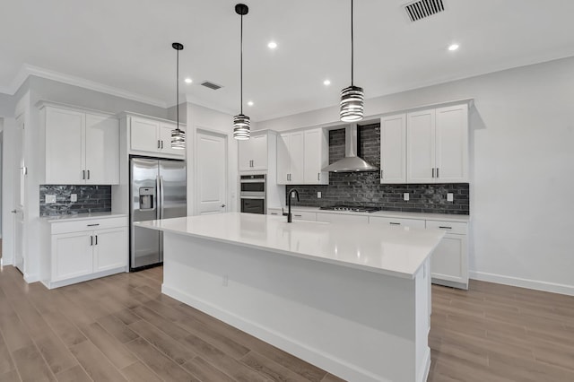 kitchen with an island with sink, appliances with stainless steel finishes, wall chimney range hood, and white cabinets