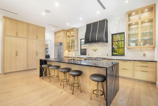 kitchen with sink, wall chimney exhaust hood, built in refrigerator, an island with sink, and light wood-type flooring