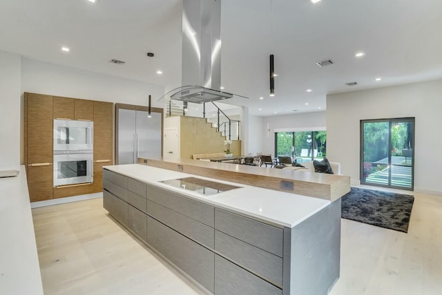 kitchen featuring a large island, multiple ovens, island exhaust hood, black electric cooktop, and light wood-type flooring