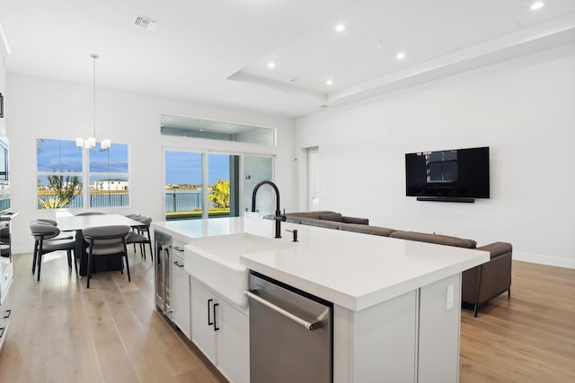 kitchen featuring white cabinetry, hanging light fixtures, stainless steel dishwasher, an island with sink, and light wood-type flooring