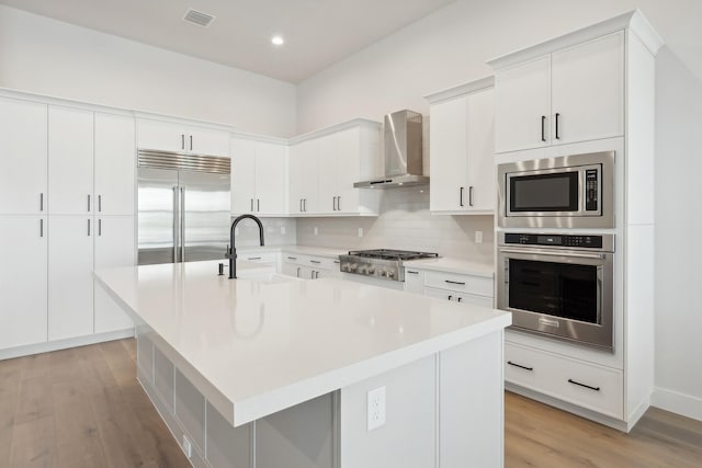 kitchen with white cabinets, built in appliances, a kitchen island with sink, and wall chimney exhaust hood