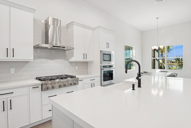 kitchen with white cabinetry, stainless steel appliances, wall chimney range hood, backsplash, and a chandelier