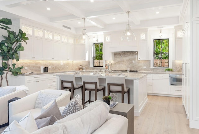 kitchen featuring coffered ceiling, white cabinets, hanging light fixtures, light wood-type flooring, and an island with sink