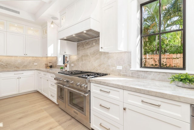 kitchen with light wood-type flooring, premium range hood, light stone counters, range with two ovens, and white cabinetry