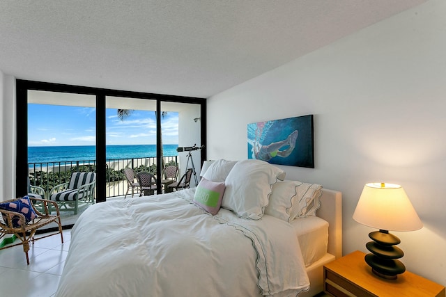 tiled bedroom featuring a wall of windows, a view of the beach, a textured ceiling, access to outside, and a water view