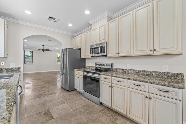 kitchen featuring cream cabinets, crown molding, ceiling fan, dark stone countertops, and appliances with stainless steel finishes