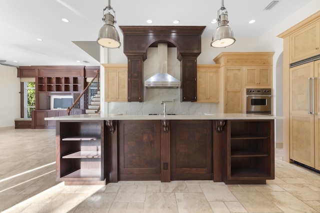 kitchen featuring light brown cabinets, wall chimney range hood, a large island with sink, pendant lighting, and decorative backsplash