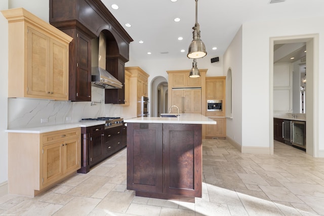 kitchen with pendant lighting, a center island with sink, built in appliances, decorative backsplash, and wall chimney exhaust hood