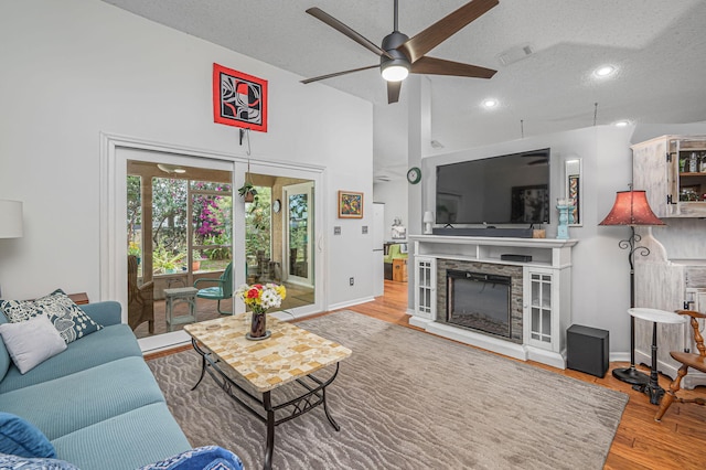 living room featuring ceiling fan, high vaulted ceiling, a textured ceiling, a fireplace, and light wood-type flooring