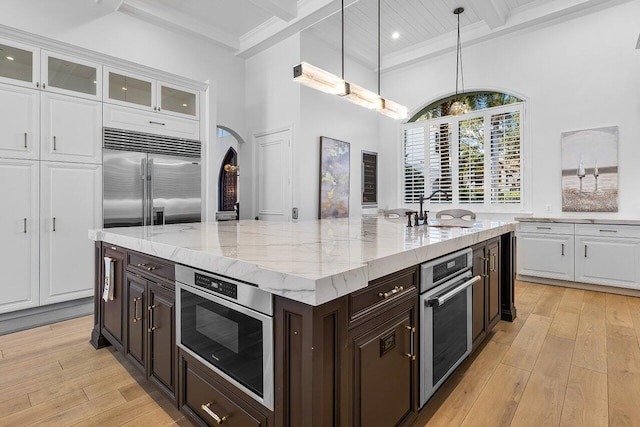 kitchen featuring appliances with stainless steel finishes, light wood-type flooring, a kitchen island with sink, decorative light fixtures, and white cabinetry