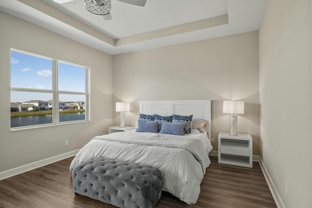 bedroom featuring ceiling fan, a water view, dark wood-type flooring, and a tray ceiling