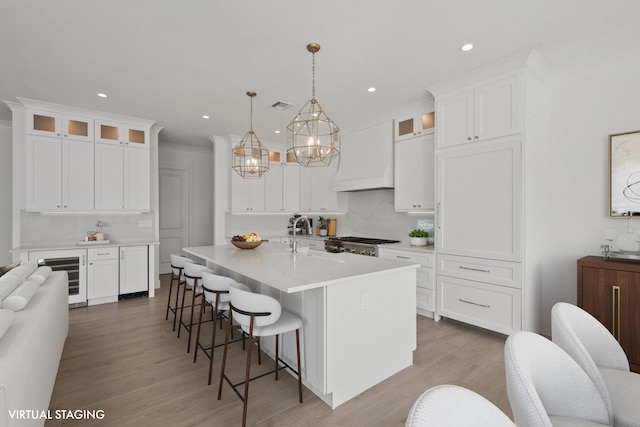 kitchen featuring an island with sink, glass insert cabinets, hanging light fixtures, custom exhaust hood, and light countertops