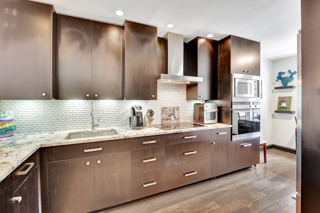 kitchen with light wood-type flooring, light stone counters, stainless steel appliances, sink, and wall chimney range hood