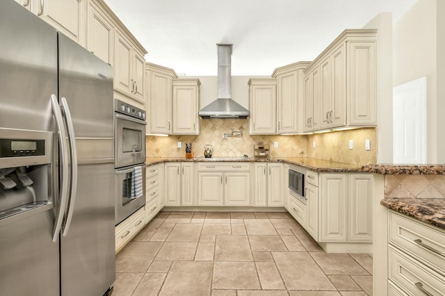 kitchen with cream cabinetry, stainless steel appliances, dark stone counters, wall chimney exhaust hood, and tasteful backsplash