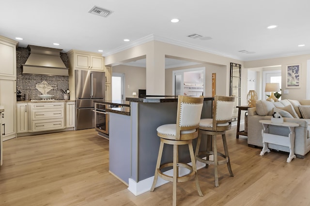 kitchen with tasteful backsplash, custom range hood, stainless steel appliances, light hardwood / wood-style floors, and cream cabinetry