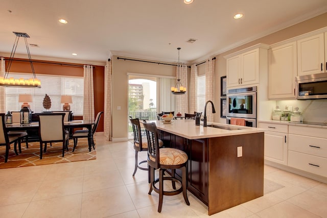 kitchen with sink, ornamental molding, an island with sink, appliances with stainless steel finishes, and decorative light fixtures