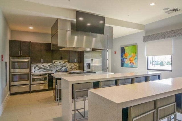 kitchen with a kitchen breakfast bar, dark brown cabinets, stainless steel appliances, wall chimney range hood, and a large island