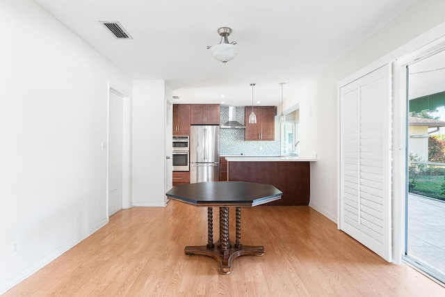 kitchen with wall chimney exhaust hood, hanging light fixtures, stainless steel appliances, light hardwood / wood-style floors, and backsplash