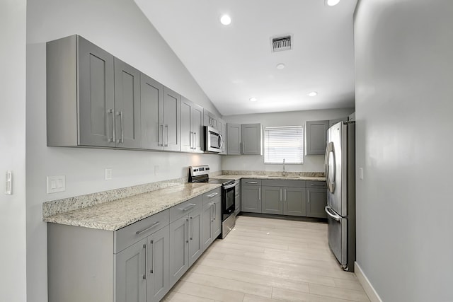 kitchen with gray cabinetry, light hardwood / wood-style flooring, vaulted ceiling, light stone countertops, and appliances with stainless steel finishes