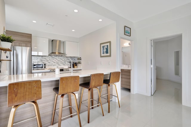 kitchen featuring white cabinets, a center island with sink, wall chimney range hood, high end fridge, and a kitchen bar