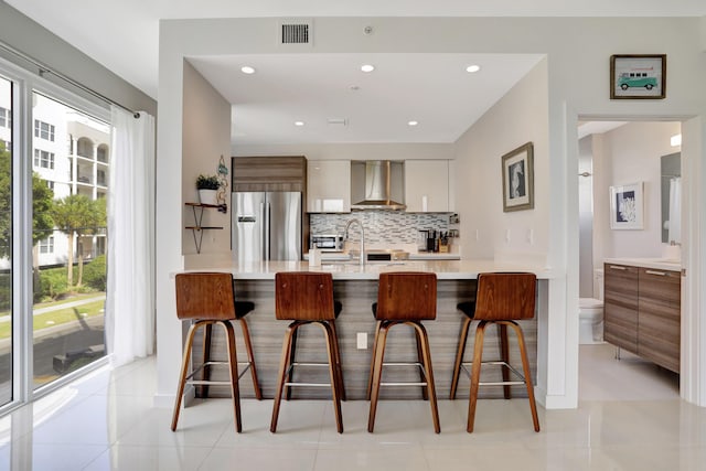 kitchen featuring stainless steel refrigerator with ice dispenser, wall chimney exhaust hood, light tile patterned flooring, a kitchen bar, and kitchen peninsula