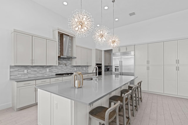 kitchen featuring wall chimney exhaust hood, stainless steel appliances, a center island with sink, an inviting chandelier, and sink