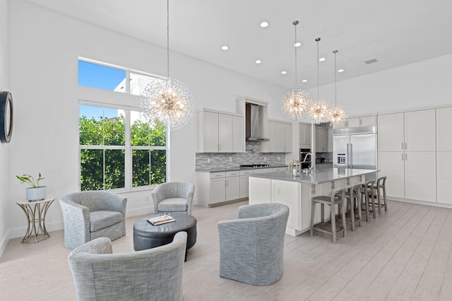 kitchen featuring a kitchen island with sink, appliances with stainless steel finishes, hanging light fixtures, a notable chandelier, and wall chimney range hood