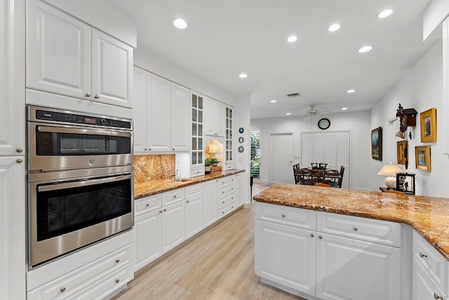 kitchen with light stone counters, double oven, ceiling fan, white cabinets, and light hardwood / wood-style floors