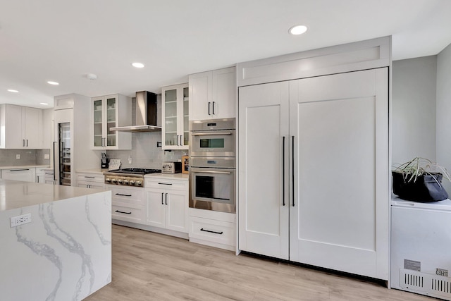 kitchen featuring appliances with stainless steel finishes, backsplash, light stone counters, wall chimney exhaust hood, and white cabinetry