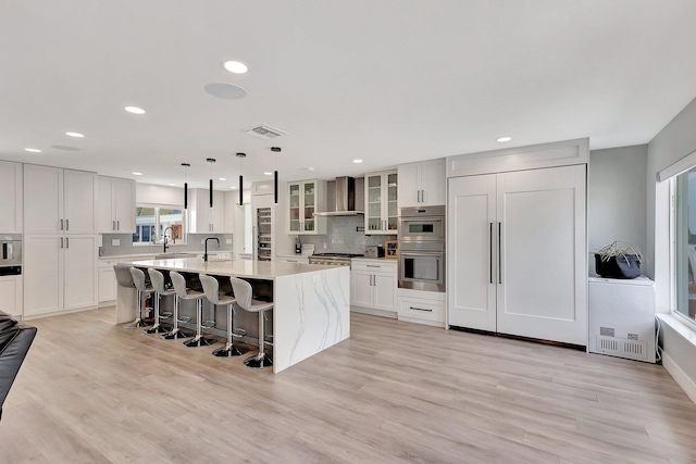 kitchen featuring wall chimney range hood, decorative light fixtures, light hardwood / wood-style floors, a breakfast bar area, and an island with sink