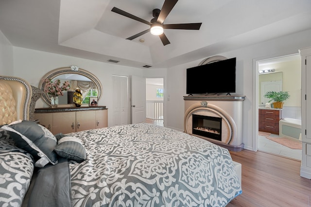 bedroom featuring light hardwood / wood-style floors, ensuite bath, ceiling fan, and a tray ceiling