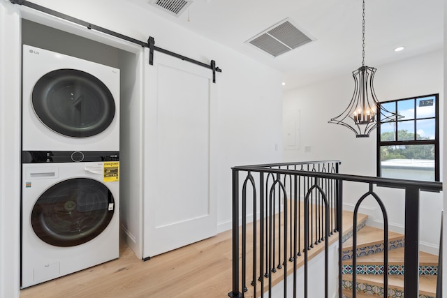 clothes washing area featuring light wood-type flooring, stacked washer / drying machine, a barn door, and a notable chandelier