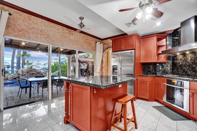 kitchen featuring stainless steel appliances, a kitchen island, dark stone counters, wall chimney range hood, and a kitchen breakfast bar