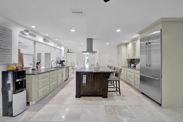 kitchen featuring built in fridge, dark stone counters, island range hood, and cream cabinetry