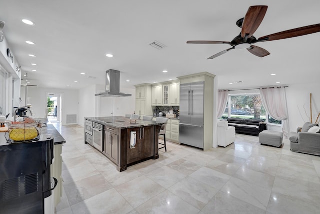 kitchen with ceiling fan, a center island, wall chimney range hood, dark stone counters, and a kitchen bar