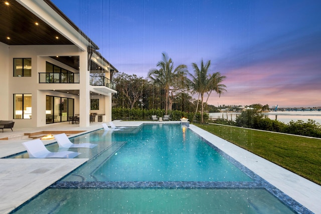 pool at dusk with a water view, a patio area, and a lawn