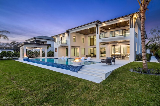 back house at dusk featuring a patio area, a balcony, a yard, an outdoor fire pit, and ceiling fan