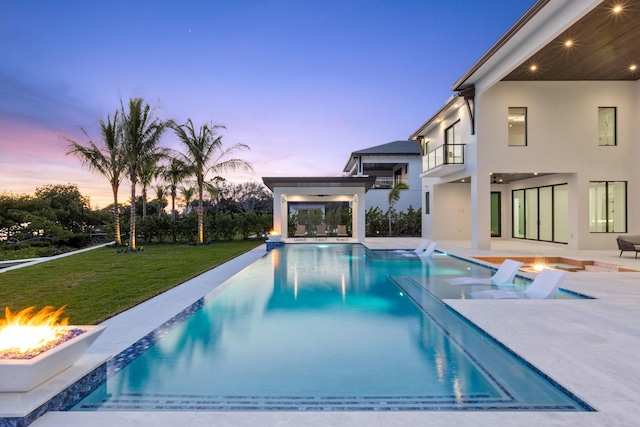 pool at dusk with a patio area, a fire pit, and a lawn