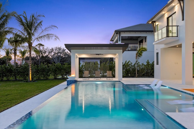 pool at dusk featuring a patio area, ceiling fan, and a lawn