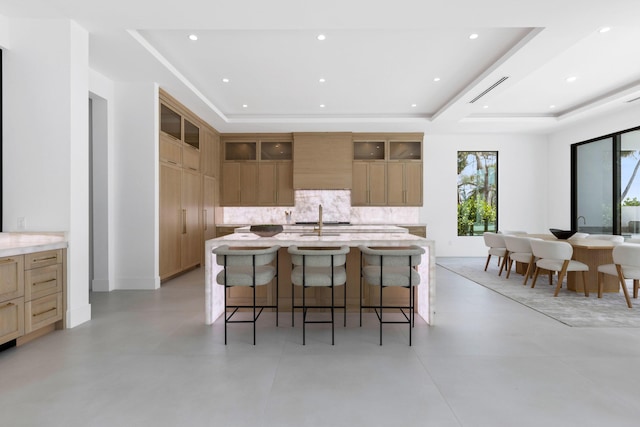 kitchen featuring sink, tasteful backsplash, a kitchen breakfast bar, a raised ceiling, and an island with sink